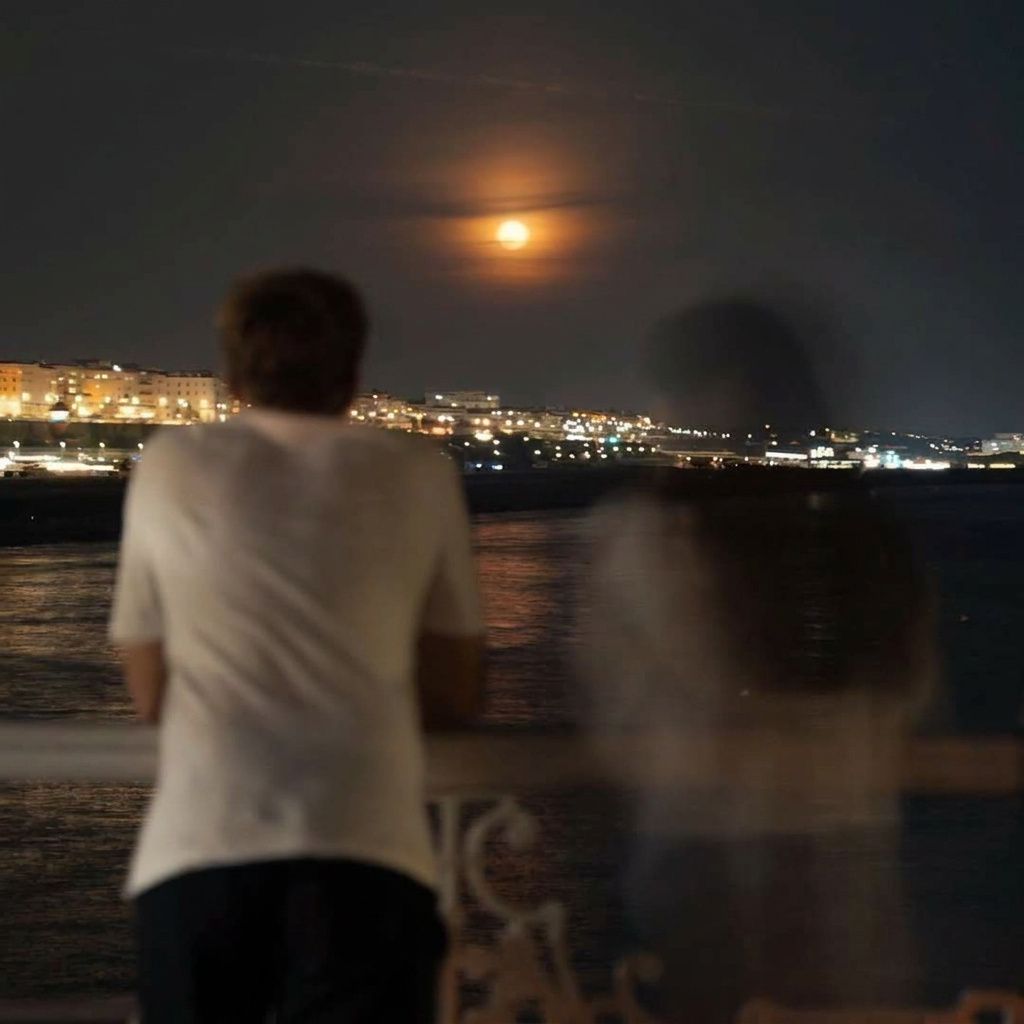 A man stands at a waterfront railing at night beside the blurred, ghostlike figure of a woman fading away